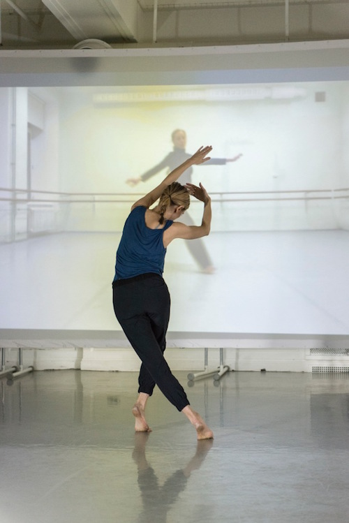 A blonde woman's back is towards us. Her left leg cross behind her while her left arm reaches above her head. Another woman is projected on screen with her arms extended out from her sides.